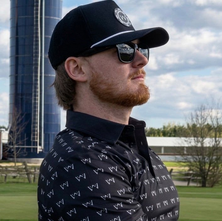 Man wearing wedge and wood black poker hat and og polo shirt with sunglasses. There is a silo and farm buildings in the background