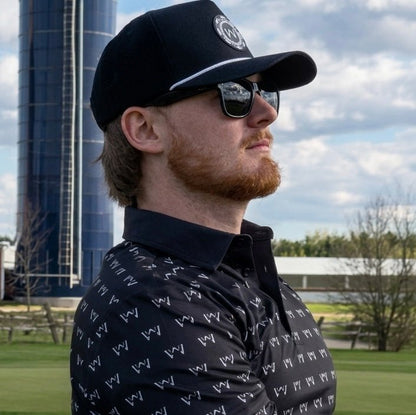 Man wearing wedge and wood black poker hat and og polo shirt with sunglasses. There is a silo and farm buildings in the background